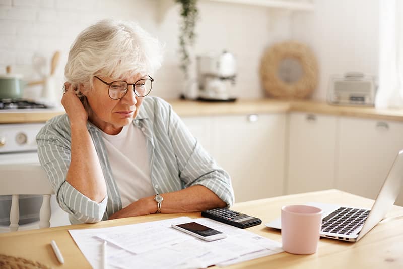 People, age, technology and finances. Depressed unhappy retired woman paying domestic bills online, trying hard to make both ends meet, sitting at kitchen table, surrounded with papers, using gadgets